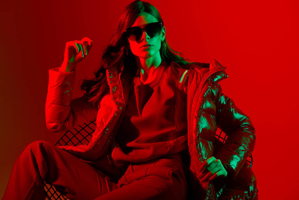Fashion portrait of elegant trendy woman in red down jacket. Neon light, sunglasses, studio shot - Foto, imagen