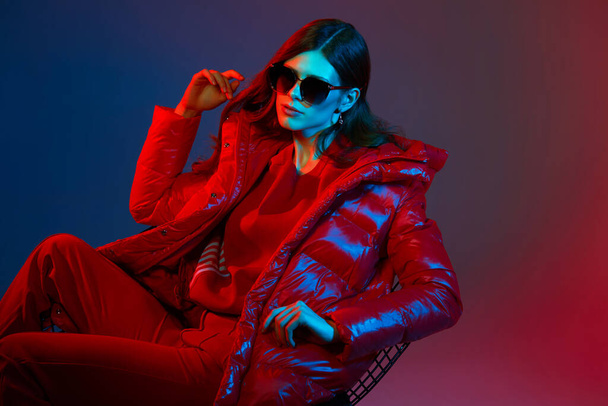 Fashion portrait of elegant trendy woman in red down jacket. Neon light, sunglasses, studio shot - Valokuva, kuva
