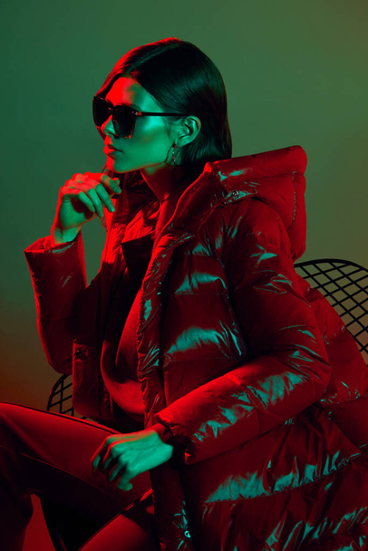 Fashion portrait of elegant trendy woman in red down jacket. Neon light, sunglasses, studio shot - Photo, image