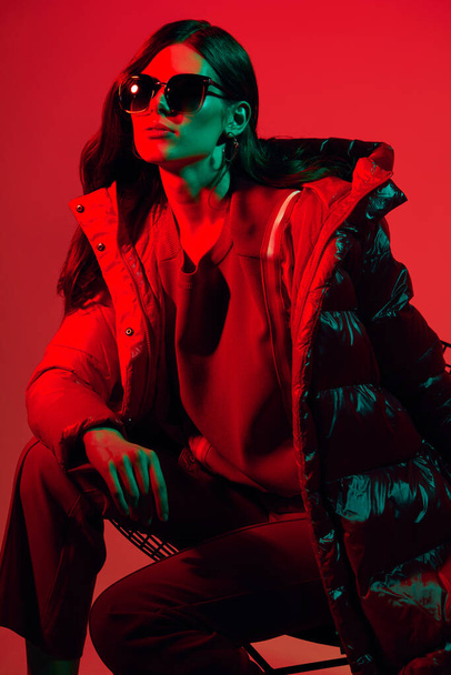 Fashion portrait of elegant trendy woman in red down jacket. Neon light, sunglasses, studio shot - Φωτογραφία, εικόνα