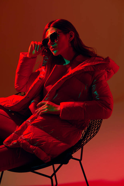 Fashion portrait of elegant trendy woman in red down jacket. Neon light, sunglasses, studio shot - Fotografie, Obrázek