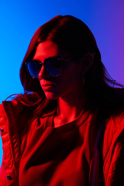Fashion portrait of elegant trendy woman in red down jacket. Neon light, sunglasses, studio shot - Foto, Bild
