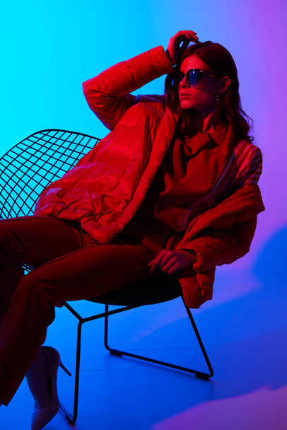 Fashion portrait of elegant trendy woman in red down jacket. Neon light, sunglasses, studio shot - Fotoğraf, Görsel