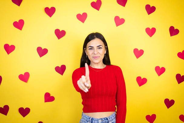 Jonge blanke vrouw over gele achtergrond met rode harten tonen en wijzen met vingers nummer een terwijl is ernstig - Foto, afbeelding