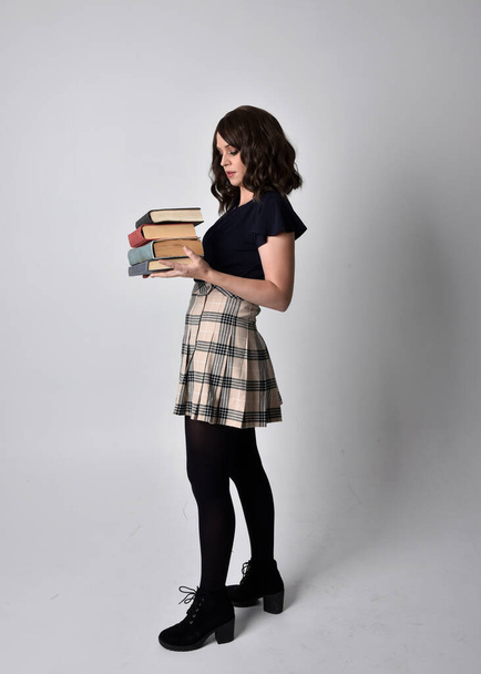 Ganzkörperporträt einer hübschen brünetten Frau, die Schottenrock und Stiefel trägt. Stehende Pose mit Büchern vor Studiohintergrund. - Foto, Bild