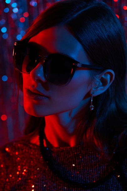 Fashion portrait of young elegant trendy woman in sunglasses. Neon light. Colored glitter tinsel background - 写真・画像