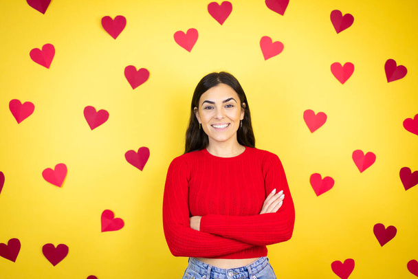 Jonge blanke vrouw over gele achtergrond met rode harten met een vrolijk gezicht staande en lachend met een zelfverzekerde glimlach tanden met gekruiste armen - Foto, afbeelding