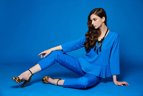 High fashion portrait of young elegant woman. Blue blouse, blue pants, curly hair, leopard shoes. Blue background - 写真・画像