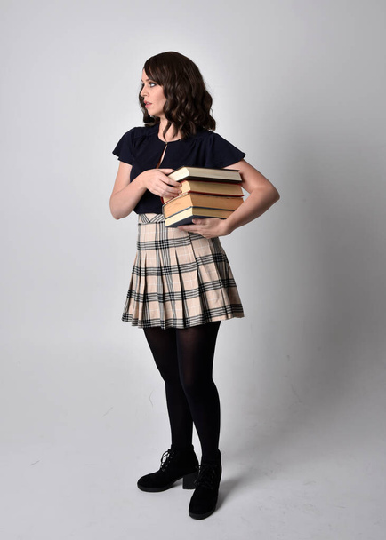 Portret pięknej brunetki w tartanowej spódnicy i butach. Stojąca pozycja do trzymania książek na tle studia. - Zdjęcie, obraz