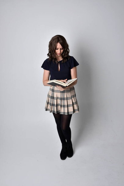 Ganzkörperporträt einer hübschen brünetten Frau, die Schottenrock und Stiefel trägt. Stehende Pose mit Büchern vor Studiohintergrund. - Foto, Bild