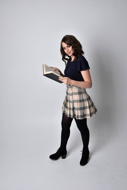 Portret pięknej brunetki w tartanowej spódnicy i butach. Stojąca pozycja do trzymania książek na tle studia. - Zdjęcie, obraz