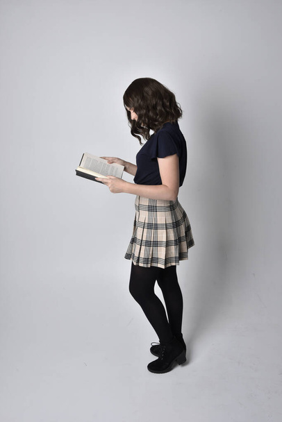 full length portrait of pretty brunette woman wearing tartan skirt and boots.  Standing pose holding books against a  studio background. - Photo, Image
