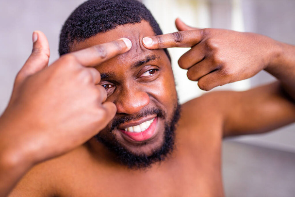african american man looking at himself in the bathroom mirror squeezing pimple - Photo, Image