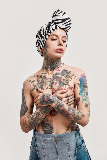Young pierced and tattooed woman standing topless with a turban over her head covering her breasts with her hands crossed on her chest - Zdjęcie, obraz
