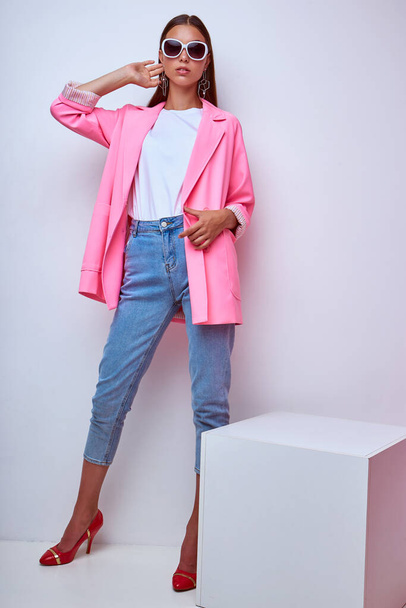 fashion portrait of young elegant woman. Pink jacket, blue jeans, sunglasses. Studio shot - 写真・画像