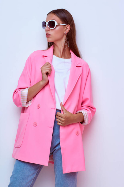 fashion portrait of young elegant woman. Pink jacket, blue jeans, sunglasses. Studio shot - Photo, Image