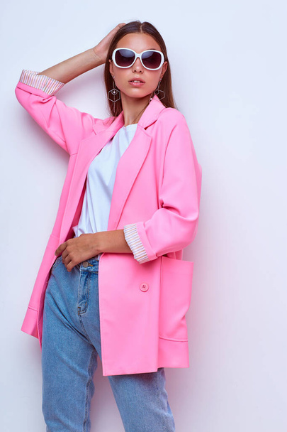 fashion portrait of young elegant woman. Pink jacket, blue jeans, sunglasses. Studio shot - Zdjęcie, obraz