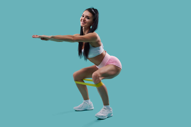 Pełna długość młodej i szczupłej uśmiechniętej kobiety w sportowej odzieży ćwiczącej z opaską oporową na niebieskim tle - Zdjęcie, obraz