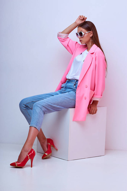 fashion portrait of young elegant woman. Pink jacket, blue jeans, sunglasses. Studio shot - Photo, Image
