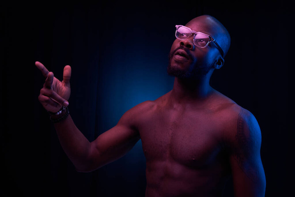 portrait of a handsome black man in red and blue light on dark background  with glasses on his eyes, he has naked sports torso and bracelets on arms - Foto, Bild