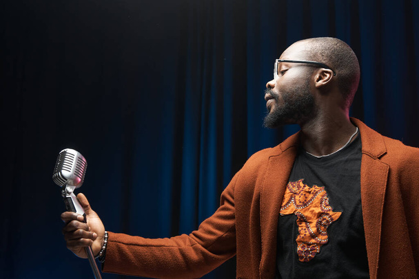portrait of a dark-skinned handsome guy in brown jacket and black t-shirt stands with a microphone and emotionally sings in a studio on dark background - Fotó, kép