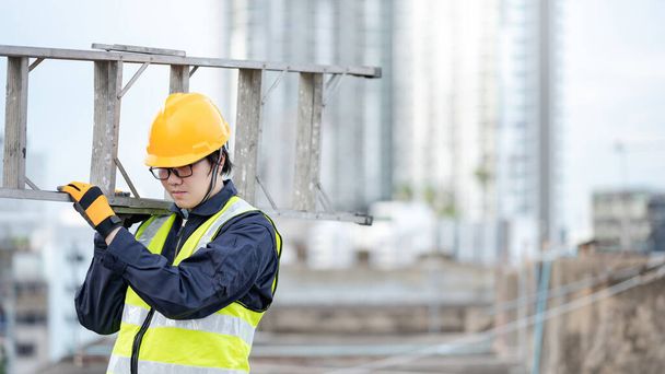 Ein asiatischer Wartungsarbeiter mit Schutzhelm und reflektierendem Anzug trägt auf der Baustelle eine Leiter aus Aluminium. Bauingenieurwesen, Architekturbau- und Haustechnikkonzepte - Foto, Bild