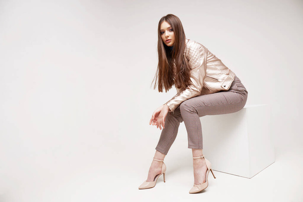 Fashion model in golden leather jacket posing in studio. Brunette hair, brown pants, shoes - Photo, image