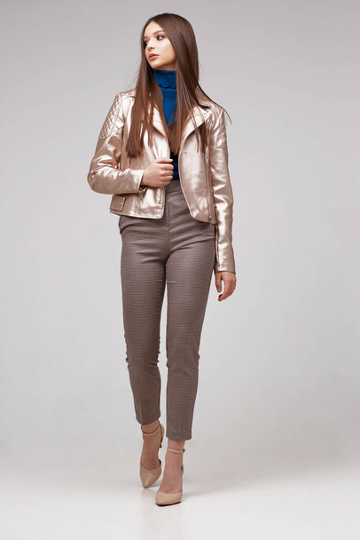 Fashion model in golden leather jacket posing in studio. Brunette hair, brown pants, shoes - Photo, Image