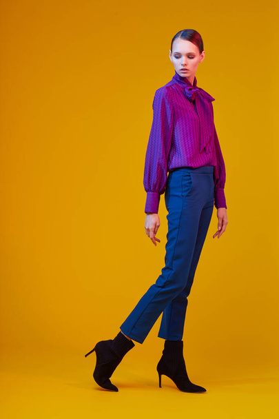 High fashion portrait of young elegant woman. Sudio shot. Violet blouse, blue pants, ankle boots, yellow background - Photo, Image