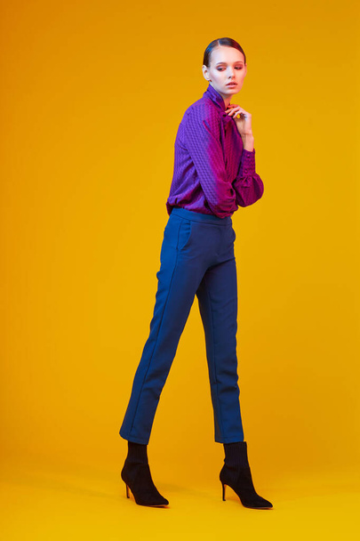 High fashion portrait of young elegant woman. Sudio shot. Violet blouse, blue pants, ankle boots, yellow background - Photo, image