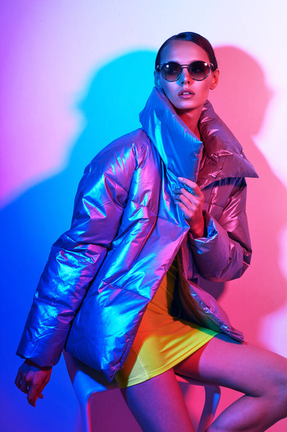 Fashion portrait of young elegant woman in sunglasses. Silver down jacket, neon light - Foto, Imagen