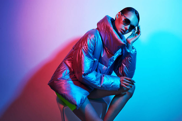Fashion portrait of young elegant woman in sunglasses. Silver down jacket, neon light - Photo, image