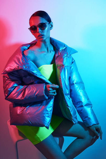 Fashion portrait of young elegant woman in sunglasses. Silver down jacket, neon light - Foto, afbeelding