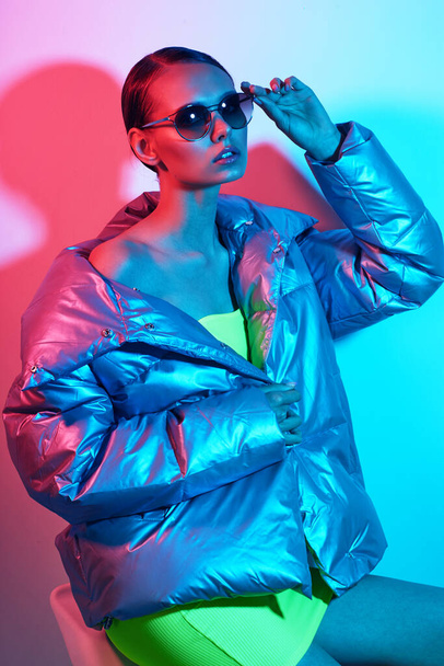 Fashion portrait of young elegant woman in sunglasses. Silver down jacket, neon light - Fotoğraf, Görsel