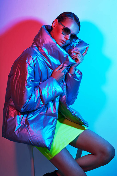 Fashion portrait of young elegant woman in sunglasses. Silver down jacket, neon light - Фото, зображення