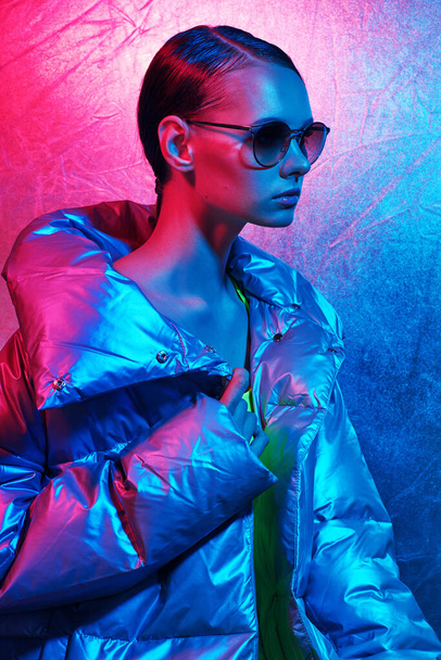 Fashion portrait of young elegant woman in sunglasses. Silver down jacket, neon light - Foto, Imagen