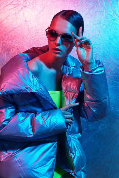 Fashion portrait of young elegant woman in sunglasses. Silver down jacket, neon light - Photo, Image