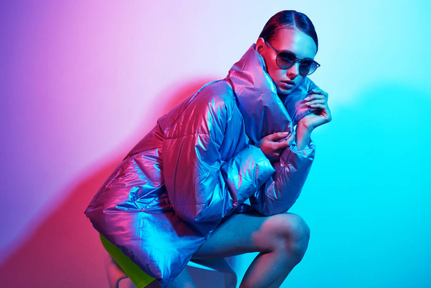 Fashion portrait of young elegant woman in sunglasses. Silver down jacket, neon light - Photo, image