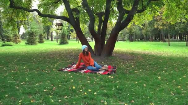 Ευτυχισμένη γυναίκα με φθινοπωρινά ρούχα κάθεται σε καρό κουβέρτα κάτω από το δέντρο στο πάρκο - Πλάνα, βίντεο