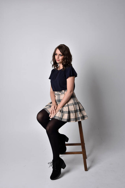 Close up portrait of pretty brunette woman wearing tartan skirt and blouse.  Sitting pose against a  studio background. - Photo, Image