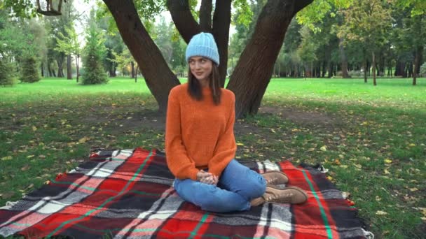 femme souriante en tenue d'automne élégant assis sur une couverture à carreaux dans le parc  - Séquence, vidéo