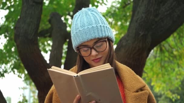 молодая женщина читает книгу и улыбается, глядя на камеру в парке - Кадры, видео
