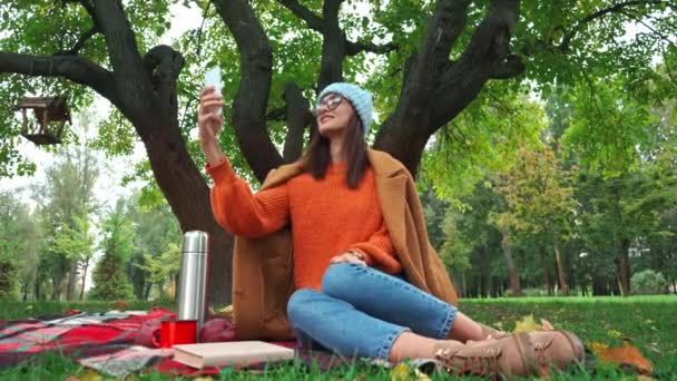 wesoła kobieta robi selfie na smartfonie na kocu w parku - Materiał filmowy, wideo