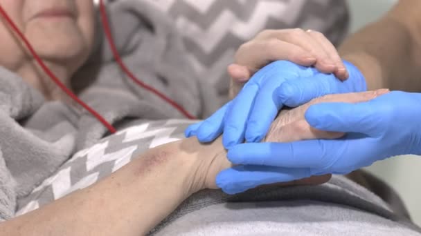 Φροντιστής κρατώντας το χέρι του ασθενούς, κατ 'οίκον φροντίδα, βοήθεια στο γηροκομείο. Ηλικιωμένη γυναίκα στο κρεβάτι με μια νοσοκόμα στο σπίτι. Κρατώντας τα χέρια μαζί. - Πλάνα, βίντεο