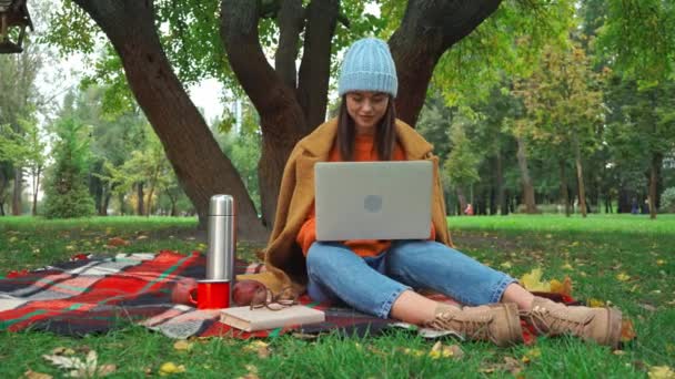 szabadúszó őszi ruhában gépelés laptopon kockás takarón a parkban - Felvétel, videó