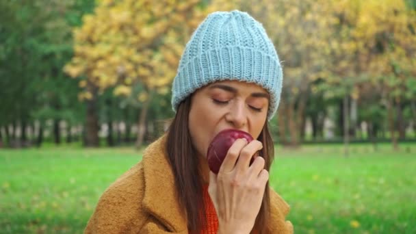 radosna kobieta jedząca soczyste jabłko patrząc na kamerę w parku - Materiał filmowy, wideo