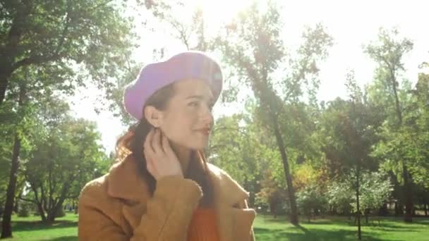 uśmiechnięta kobieta w jesiennych ubraniach dotykająca włosów podczas spaceru po parku - Materiał filmowy, wideo