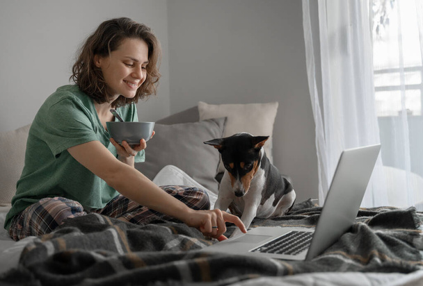 Счастливая молодая женщина у себя дома завтракает на кровати в пижаме перед ноутбуком с африканским терьером бассенджи - Фото, изображение