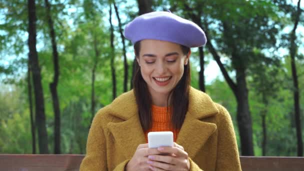 vrolijke vrouw in stijlvolle herfst outfit chatten op smartphone in park - Video