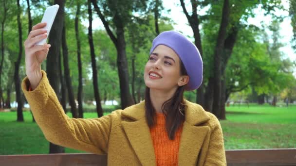 スタイリッシュな秋の服を着た幸せな女性が公園でスマートフォンで自撮り - 映像、動画